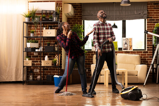 African american couple enjoying spring cleaning by turning household chores into music time. Black woman pretends mop is microphone as male partner sings, creating joyful dance number in apartment.