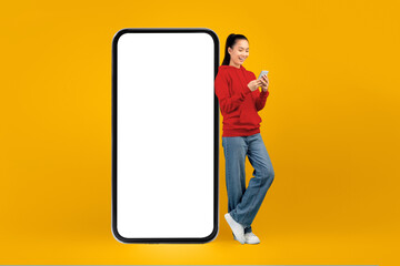 A young woman stands next to a large blank phone display. She is looking at her smartphone and smiling. The background is bright yellow, creating a lively atmosphere. © Prostock-studio