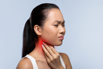 A woman stands in an indoor space, holding her neck with a pained expression. She appears to be experiencing discomfort or pain in her throat area, possibly due to an illness. © Prostock-studio