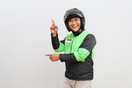 Excited and smiling Asian online motorbike taxi driver looking at camera while pointing sideways in empty space, isolated on grey background.