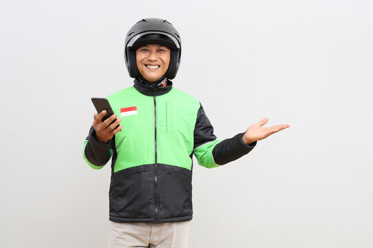 Friendly Asian online motorbike taxi driver wearing green jacket standing holding mobile phone and presenting something beside in empty space for advertisement, promotion or text.
