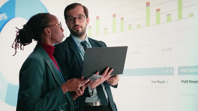 Female executive and business partner compile shareholder financial report using laptop in conference room. African american woman and coworker look at key performance indicators earnings data