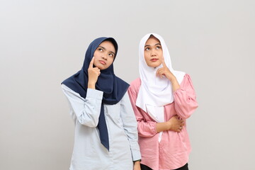 Fototapeta premium Two young Muslim women in hijab posing and looking up, contemplating or thinking together looking for an idea or solution, isolated on a grey background.