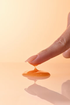 Finger touching a drop of amber liquid against a soft, warm background