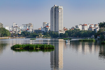 Fototapeta premium A scenic view of Hanoi, Vietnam, showcasing the serene beauty of Truc Bạch Lake. People enjoy swan paddle boats on the lake, with city buildings in the background.