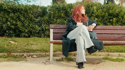Fototapeta premium Red-Haired Woman Relaxing on Park Bench with Coffee