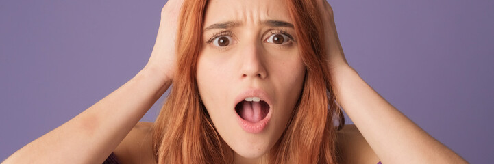 Close-up of dissatisfied red-haired woman, looks frustrated with her hands on her head on purple background in studio. Panoramic
