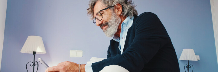 Stylish good looking mature man is sitting on bed and using smartphone. Panoramic