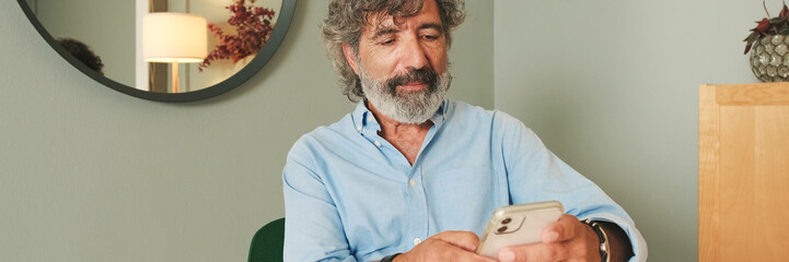 An elderly man with gray hair, wearing blue shirt, uses mobile phone while sitting in the living room in modern apartment. Panoramic