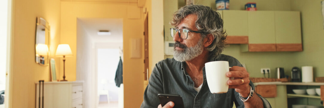 Elderly man drinks coffee from mug and uses mobile phone at home. Panoramic
