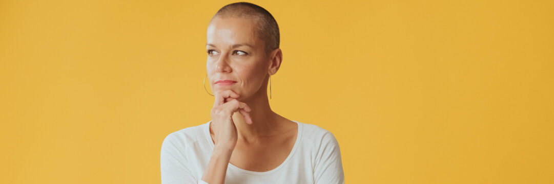 Young hairless woman, thinks over problem, remembering something, looking at camera isolated on yellow background in studio. Panoramic. Panoramic