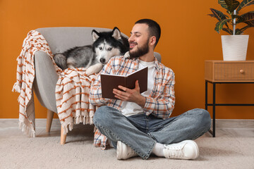 Handsome young man sitting on floor and reading book with cute husky dog at home © Pixel-Shot