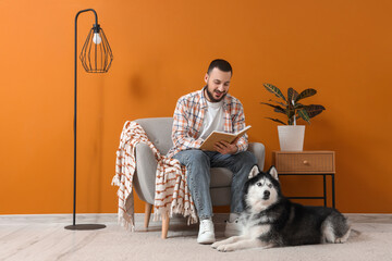 Handsome young man sitting on armchair and reading book with cute husky dog at home © Pixel-Shot