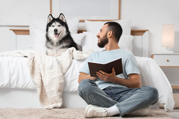 Handsome young man sitting on floor near bed and reading book with cute husky dog in bedroom © Pixel-Shot