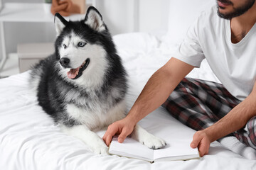 Young man sitting on bed and reading book with cute husky dog in bedroom © Pixel-Shot