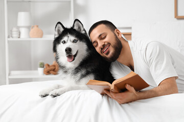 Handsome young man lying on bed and reading book with cute husky dog in bedroom © Pixel-Shot