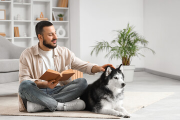 Handsome young man sitting on floor and reading book with cute husky dog in living room © Pixel-Shot