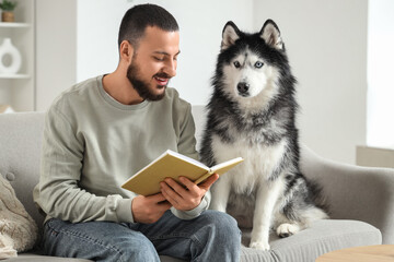 Handsome young man sitting on sofa and reading book with cute husky dog at home © Pixel-Shot