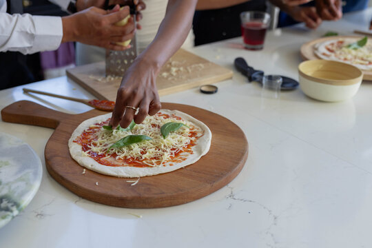 Raw pizza crust is being topped with tomato sauce, shredded cheese, basil on stone countertop