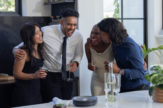 Diverse friends laughing, leaning in and holding flutes and glasses on white kitchen island