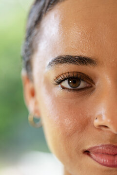 Right half of face is showing brown eye, gold hoop earring, nose stud near green foliage