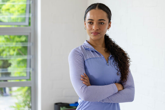 African American woman posing arms crossed in gym wearing purple zip top, window, brick wall, mats