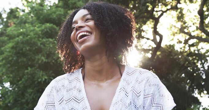 African American woman smiling laughing tilting as lens flare rising in park trees in eyelet top