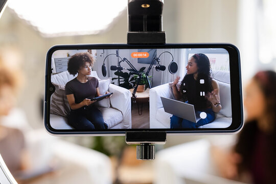 African American women hosts recording on phone screen in living room in casual clothes with mics