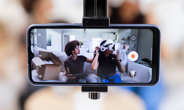 African American women friends sitting on sofa, recording on clamp-mounted phone testing VR headset