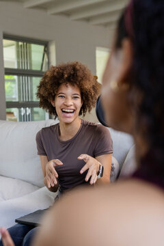 African American female friends laughing, gesturing on light sofa with closed laptop and smartwatch