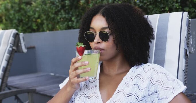 African American woman glancing at green drink on lounge at pool, lifting and sipping to cool