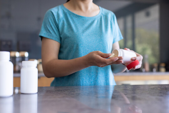 White pill bottle tipping tablet into palm on stone countertop in kitchen with two bottles