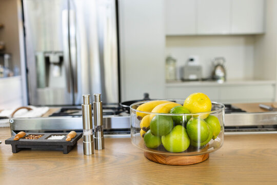 Clear glass bowl is sitting on wooden countertop island, holding limes, lemons, bananas