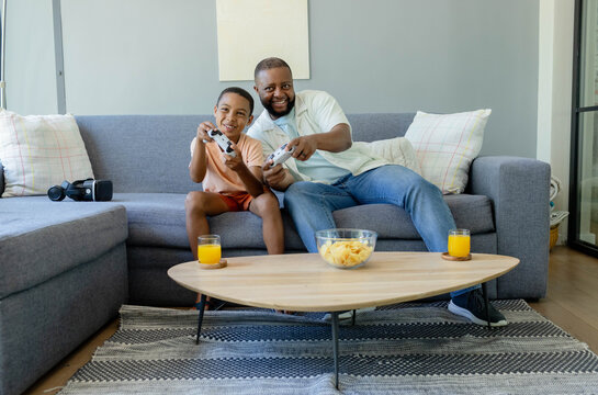 African American dad and son playing games on gray sofa at home holding white controllers