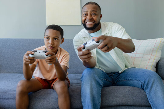 African American father and son gaming in living room, father wearing shirt holding controllers