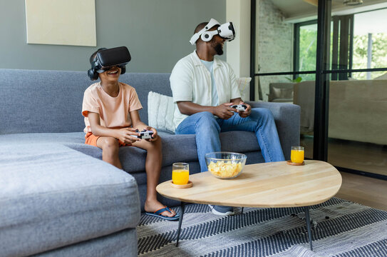 African American father and teenage son wearing VR headsets on gray sofa, holding controllers