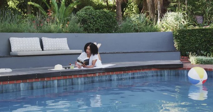 African American woman 20s lying on towel at pool reading book, sipping, glancing at floating ball