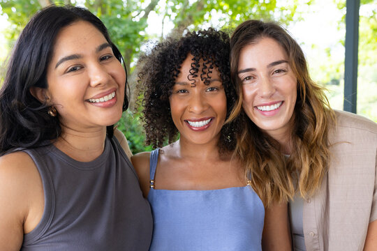 Diverse female friends smiling and posing on patio wearing blazer, tank top and earrings