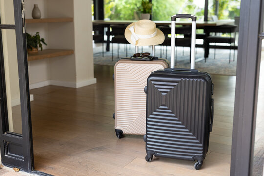 Black and beige suitcases are waiting by open doorway on light hardwood floor with hat, sunglasses