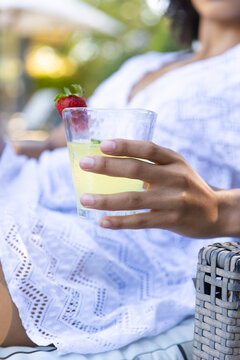 Hand is holding clear tumbler with pale yellow fizzy drink and strawberry, on wicker lounge chair