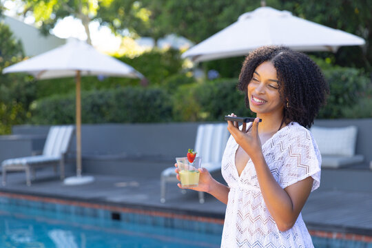 African American woman in lace cover-up on pool deck holding cocktail while using phone, copy space
