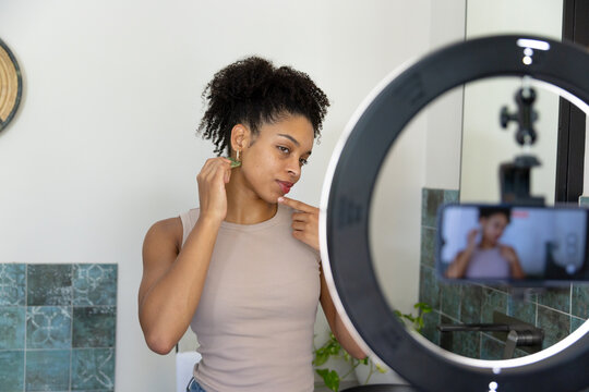 African American woman in tank top adjusting earring touching chin at sink with ring-light phone