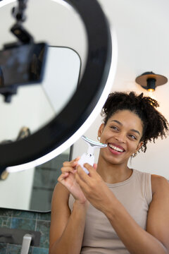 Woman standing at vanity holding T-shaped facial roller near cheek filming with ring light phone