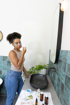 African American woman using skincare, wearing under-eye patches at bathroom sink, copy space