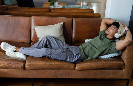 African American man lying on brown leather sofa at home in beanie and listening with headphones
