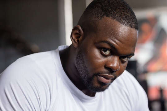 Man wearing white athletic T-shirt, perspiring and focusing toward camera in gym near metal post