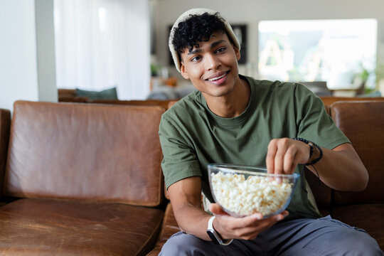 African American male sitting on leather sofa in green tee and beanie reaching into popcorn bowl