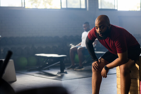 African men in gym resting now after set on plyo box near dumbbell rack, copy space