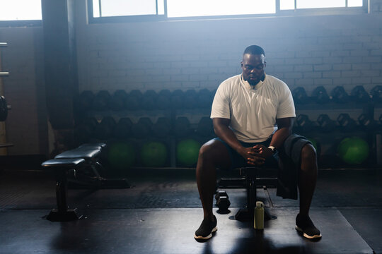 African man sitting on bench in weight room, wearing white tee, black shorts, headphones