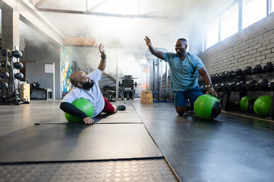 Two African American men high-fiving while exercising at gym in sportswear on green medicine balls